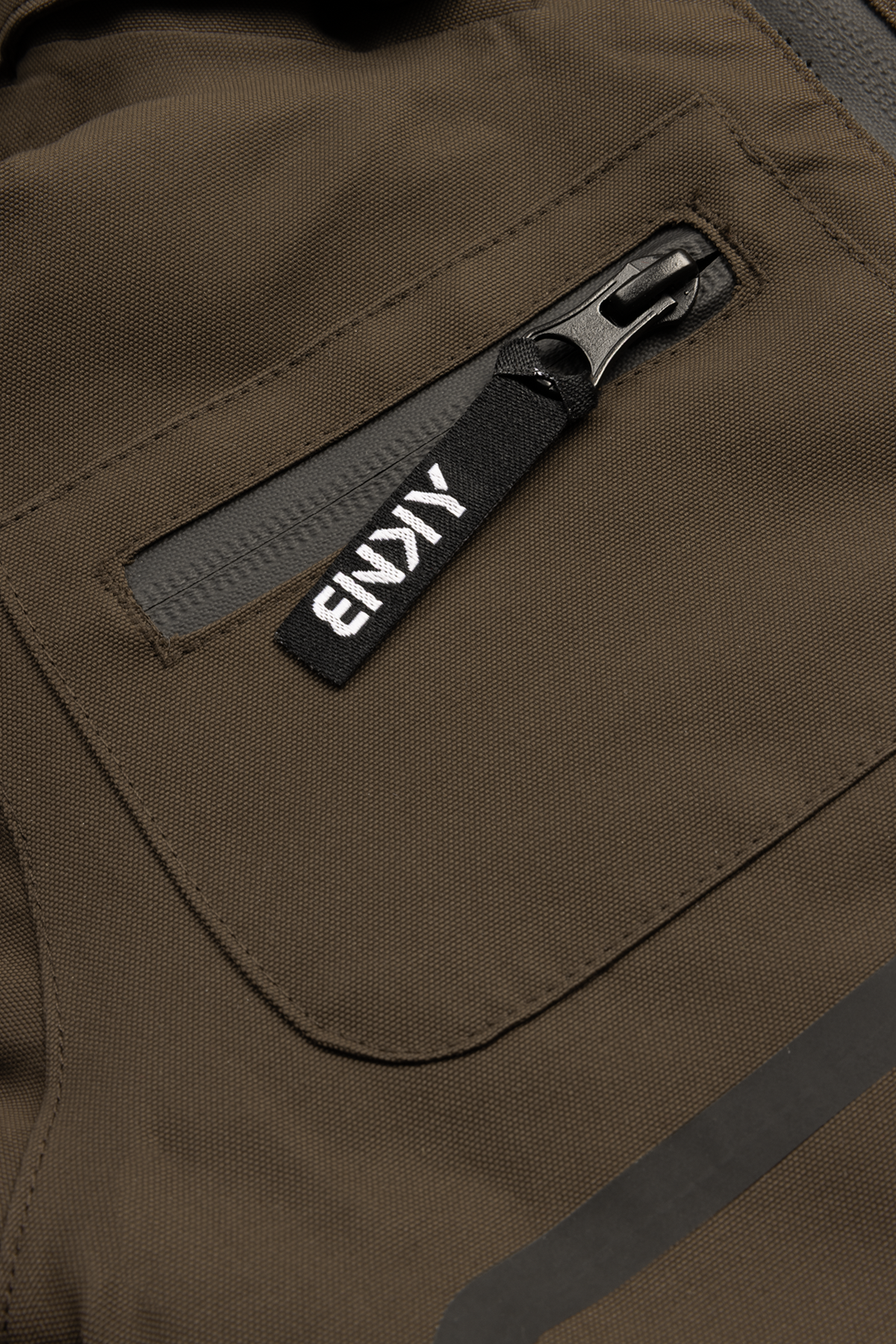 Close-up of a chocolate brown fabric featuring a zippered pocket with a black tag labeled "BNKY," showcasing stylish outdoor apparel.